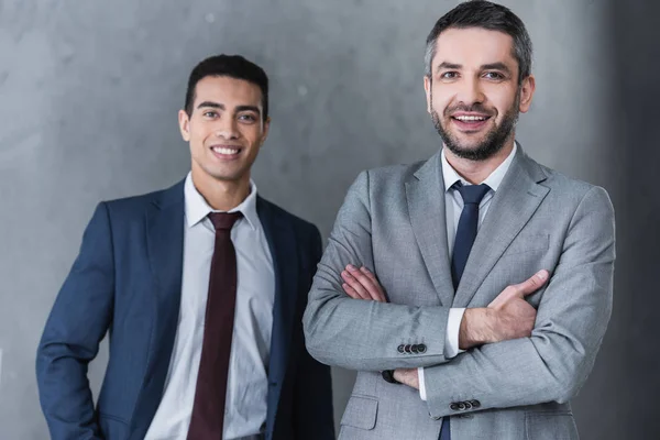 Професійні впевнені бізнесмени, що стоять разом і посміхаються на камеру на сірому — стокове фото