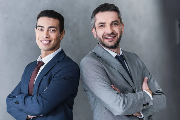 Professional confident businessmen standing with crossed arms and smiling at camera on grey — Stock Photo