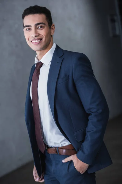 Portrait de beau jeune homme d'affaires métis debout avec la main dans la poche et souriant à la caméra — Photo de stock