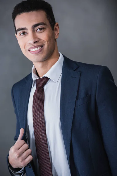 Portrait of handsome young mixed race businessman adjusting suit jacket and smiling at camera on grey — Stock Photo