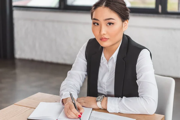 Seria asiático mujer de negocios escritura en notebook y mirando cámara en oficina - foto de stock