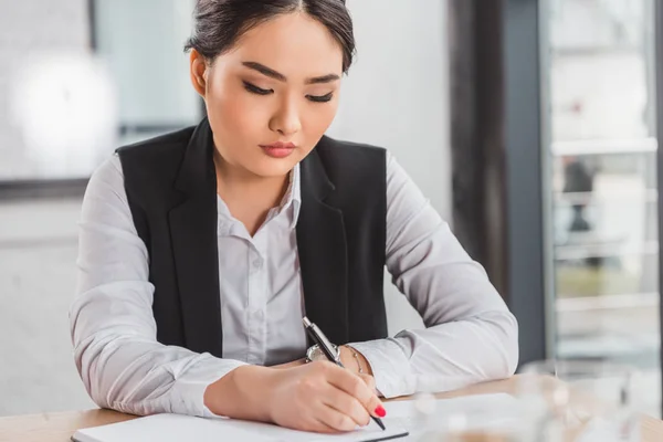 Schöne fokussierte junge kasachische Geschäftsfrau, die am Arbeitsplatz in Notizbuch schreibt — Stockfoto