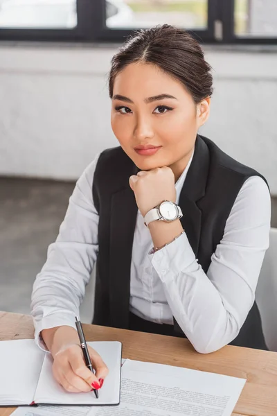 Schöne junge asiatische Geschäftsfrau sitzt mit der Hand im Kinn und lächelt in die Kamera im Büro — Stockfoto