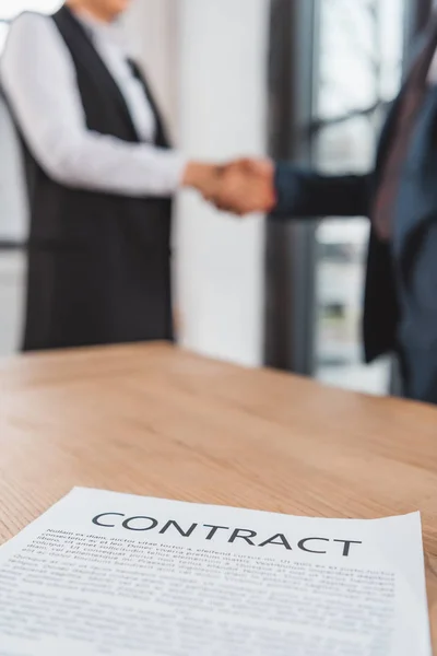 Vista de primer plano del contrato en la mesa y los empresarios estrechando la mano detrás - foto de stock