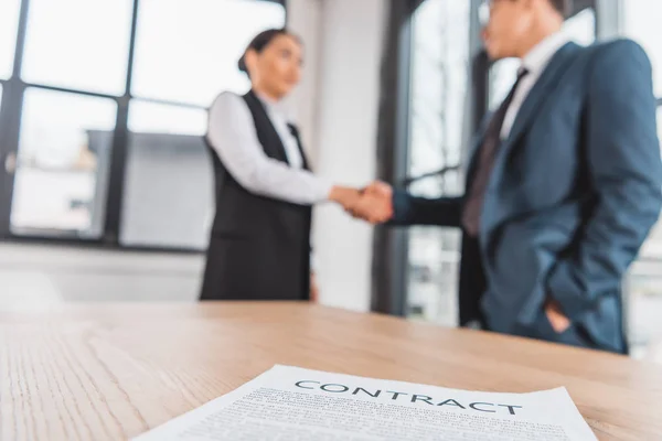 Vista de primer plano del contrato en la mesa y los empresarios estrechando la mano detrás - foto de stock