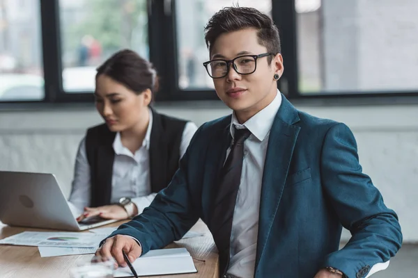 Красивый молодой азиатский бизнесмен в очках улыбается в камеру, в то время как женщина-коллега использует ноутбук в офисе — стоковое фото