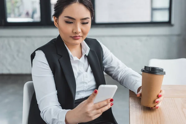 Hermosa joven empresaria sosteniendo café para ir y el uso de teléfono inteligente en la oficina - foto de stock