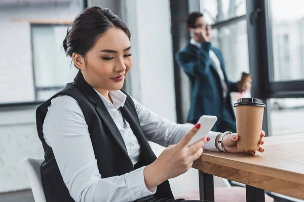 Beautiful smiling kazakh businesswoman holding coffee to go and using smartphone at workplace — Stock Photo