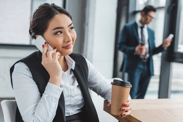 Smiling young asian businesswoman holding paper cup and talking by smartphone in office — Stock Photo