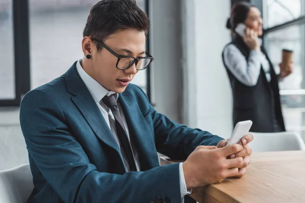 Jovem asiático empresário usando smartphone e feminino colega falando atrás no escritório — Fotografia de Stock