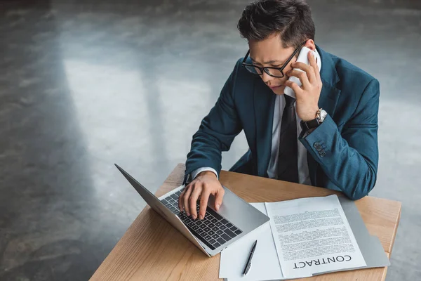 High angle view of serious young businessman in eyeglasses talking by smartphone and using laptop in office — Stock Photo