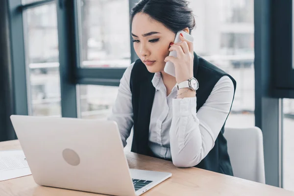 Молодая сконцентрированная деловая женщина, использующая ноутбук и разговаривающая по смартфону в офисе — стоковое фото