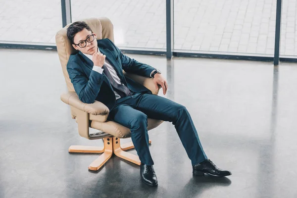 Hochwinkelaufnahme eines selbstbewussten asiatischen Geschäftsmannes mit Brille, der mit der Hand am Kinn im Büro sitzt — Stockfoto