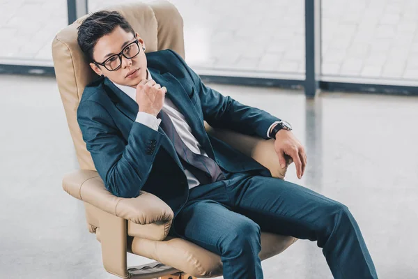 Confident asian businessman in eyeglasses sitting with hand on chin in office — Stock Photo