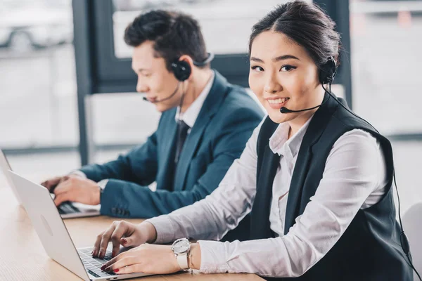 Mujer joven con auriculares sonriendo a la cámara mientras trabaja con un colega masculino en el centro de llamadas - foto de stock