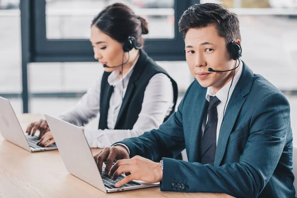 Junger Mann mit Headset blickt in Kamera, während er mit Kollegin im Callcenter arbeitet — Stockfoto