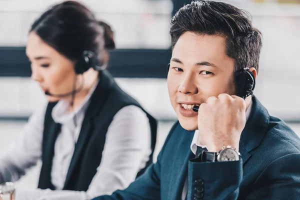 Schöner junger Mann mit Headset lächelt in die Kamera, während er mit einer Kollegin im Callcenter arbeitet — Stockfoto