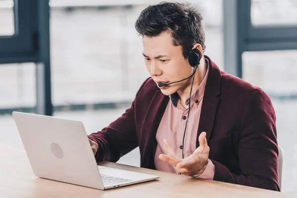 Émotionnel jeune asiatique homme d'affaires dans casque en utilisant ordinateur portable sur le lieu de travail — Photo de stock