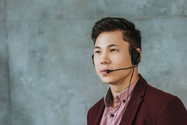 Bello giovani asiatico call center operatore guardando lontano su grigio — Foto stock