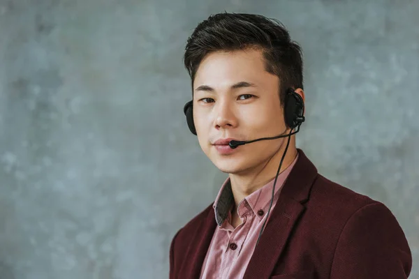 Handsome young asian call center operator looking at camera on grey — Stock Photo