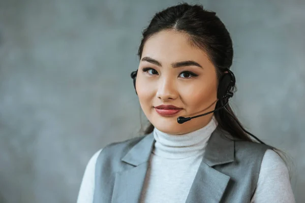 Hermoso joven operador de centro de llamadas kazakh en auriculares sonriendo a la cámara en gris - foto de stock