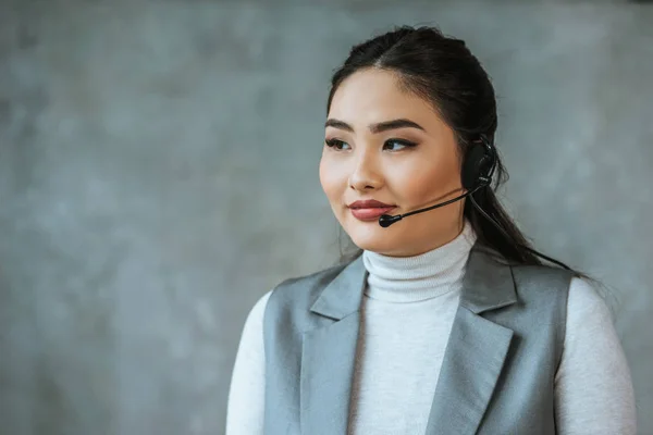 Operador de call center kazakh bonita no fone de ouvido olhando para longe em cinza — Fotografia de Stock