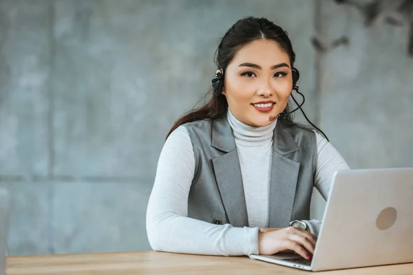 Attrayant jeune femme d'affaires dans casque de travail avec ordinateur portable et souriant à la caméra dans le bureau — Photo de stock