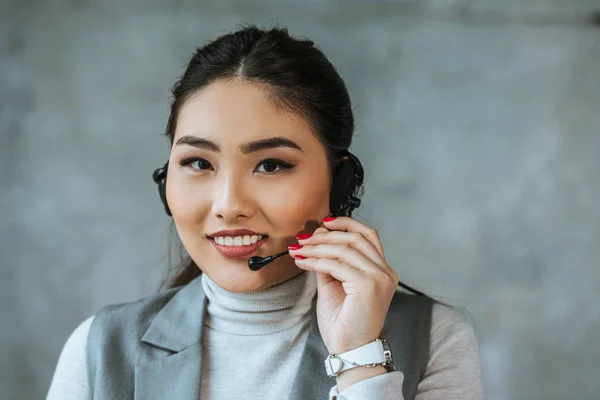 Schöne junge asiatische Callcenter-Betreiber lächelt in die Kamera auf grau — Stockfoto