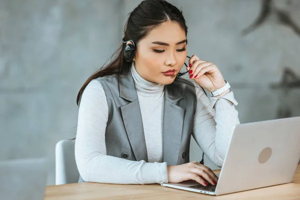 Молода азіатська бізнес-леді в гарнітурі використовує ноутбук на робочому місці — стокове фото
