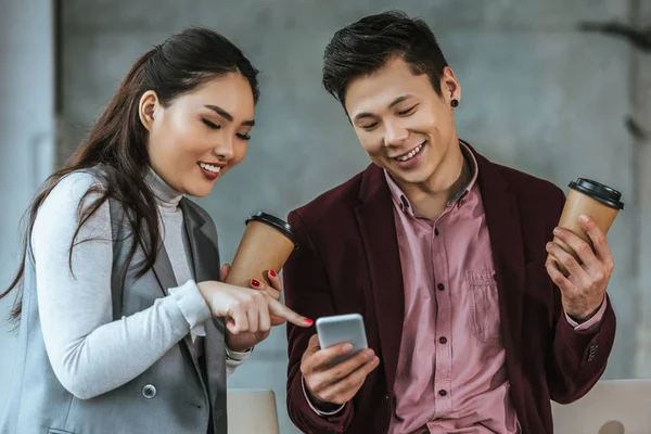 Sonrientes compañeros de trabajo jóvenes sosteniendo vasos de papel y utilizando el teléfono inteligente en la oficina - foto de stock