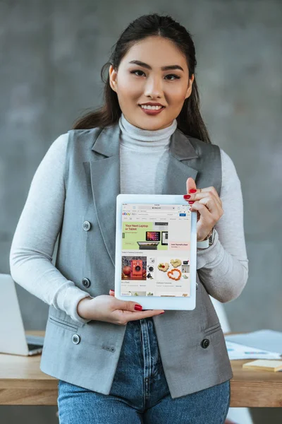 Joven asiático mujer de negocios celebración digital tablet con ebay aplicación y sonriendo a la cámara - foto de stock
