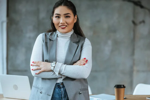 Hermosa mujer de negocios kazakh de pie con los brazos cruzados y sonriendo a la cámara en la oficina - foto de stock
