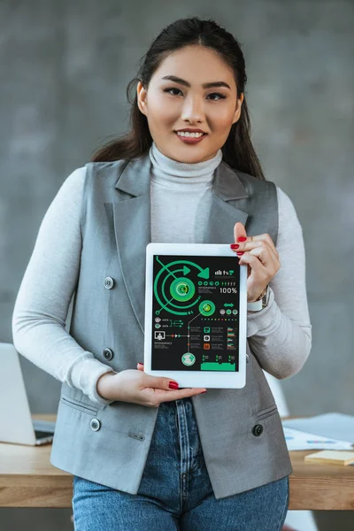 Joven asiático mujer de negocios celebración digital tableta con negocios gráficos y sonriendo a cámara - foto de stock