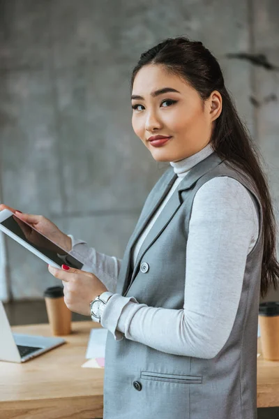 Красивая молодая азиатская бизнесвумен, использующая цифровой планшет и улыбающаяся в камеру — стоковое фото