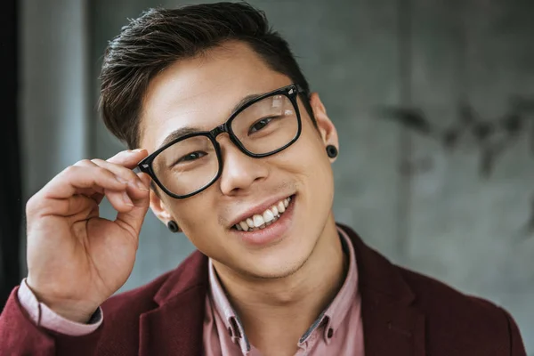 Retrato de bonito jovem asiático homem ajustando óculos e sorrindo para a câmera — Fotografia de Stock