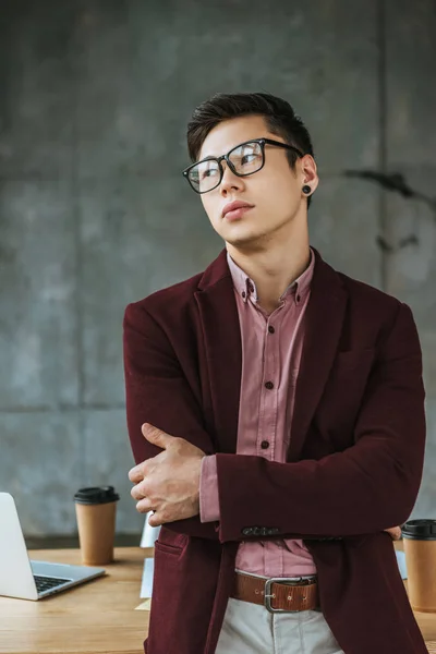 Handsome young busienssman in eyeglasses standing with crossed arms and looking away in office — Stock Photo