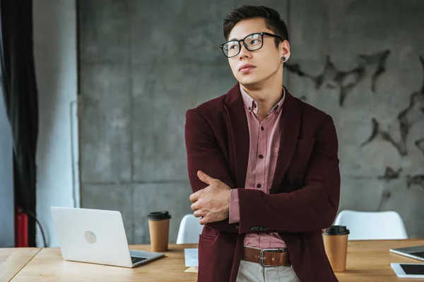 Pensivo jovem kazakh empresário em óculos de pé com braços cruzados e olhando para longe no escritório — Fotografia de Stock
