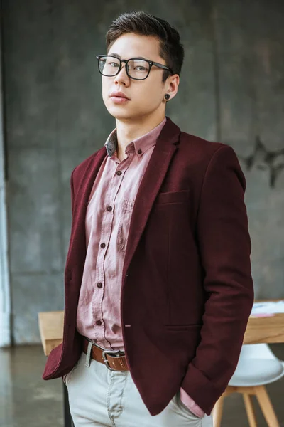 Joven guapo en gafas de pie con las manos en los bolsillos y mirando a la cámara en la oficina - foto de stock