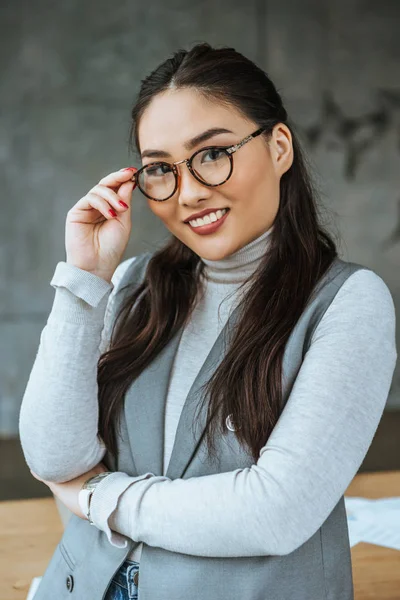 Retrato de hermosa mujer de negocios asiática ajustando gafas y sonriendo a la cámara - foto de stock