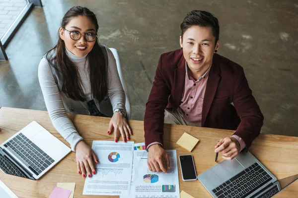 Vista ad alto angolo di felice giovani colleghi di lavoro asiatici seduti insieme e sorridente alla fotocamera in ufficio — Foto stock