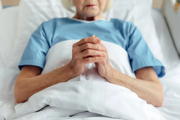 Vue recadrée de la femme âgée couchée au lit avec les mains pliées et priant à l'hôpital — Photo de stock