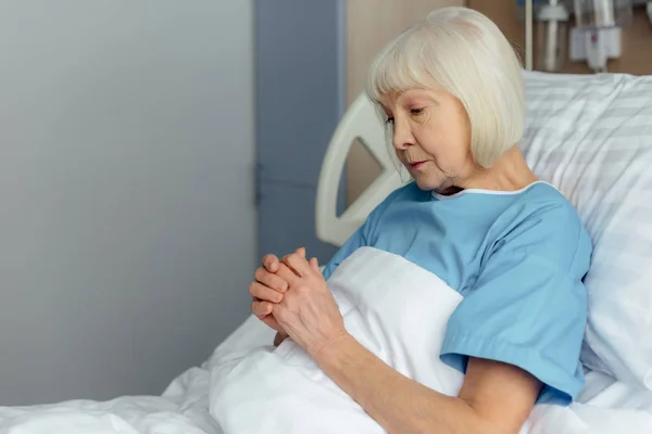 Selektiver Fokus der Seniorin, die mit gefalteten Händen im Bett liegt und im Krankenhaus betet — Stockfoto