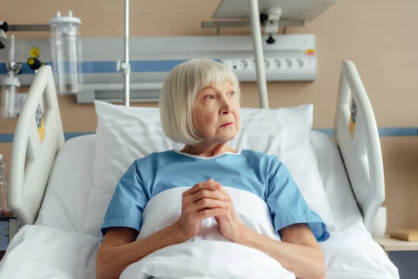 Грустная пожилая женщина лежит в постели со сложенными руками и молится в больнице — стоковое фото