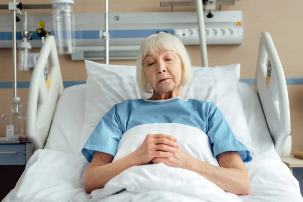 Mujer mayor con las manos dobladas acostada en la cama en el hospital - foto de stock