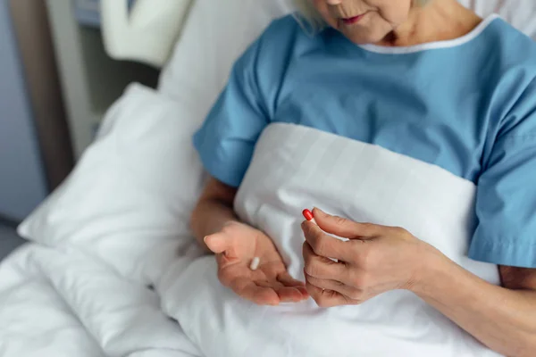 Partial view of senior woman lying in bed and holding pills in hospital — Stock Photo