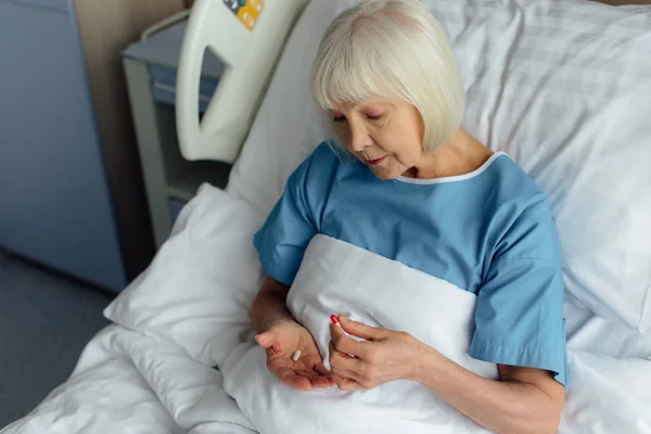 Selective focus of senior woman lying in bed and holding pills in hospital — Stock Photo