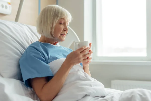 Selective focus of senior woman lying in bed and drinking tea in hospital — Stock Photo