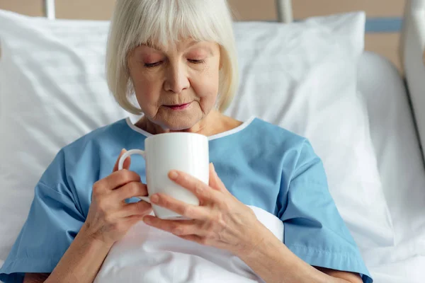Старша жінка з сірим волоссям лежить в ліжку і п'є чай в лікарні — стокове фото