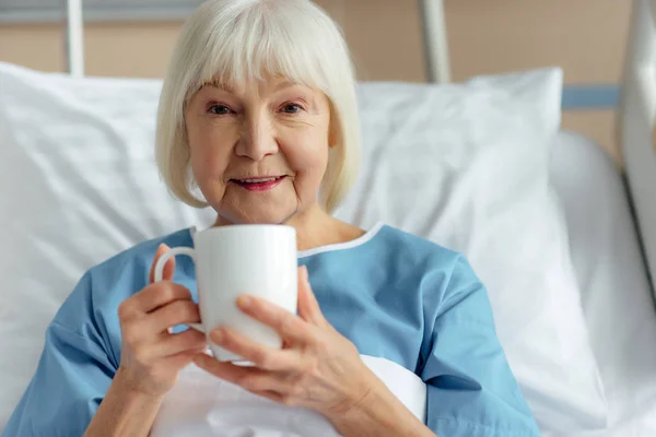 Femme âgée souriante couchée au lit, regardant la caméra et buvant du thé à l'hôpital — Photo de stock