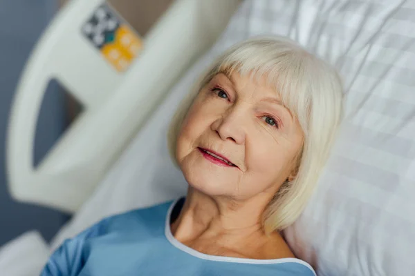 Mujer mayor sonriente con el pelo gris acostado en la cama en el hospital - foto de stock